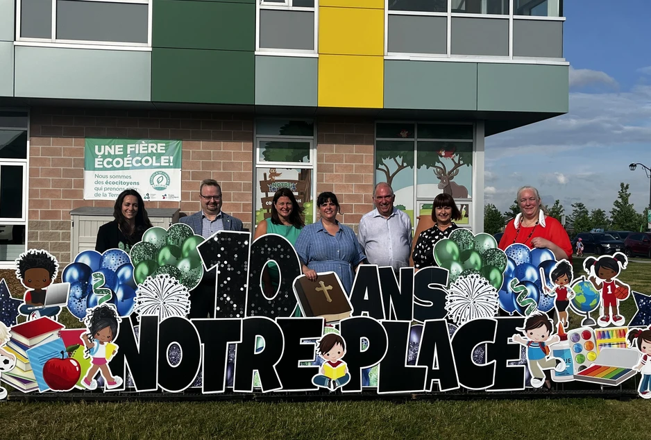 Des membres du personnel et des invités posent devant l’École élémentaire catholique Notre-Place pour souligner le 10e anniversaire de l’école. Le groupe se tient derrière une grande enseigne festive « 10 ans Notre-Place », décorée de ballons.